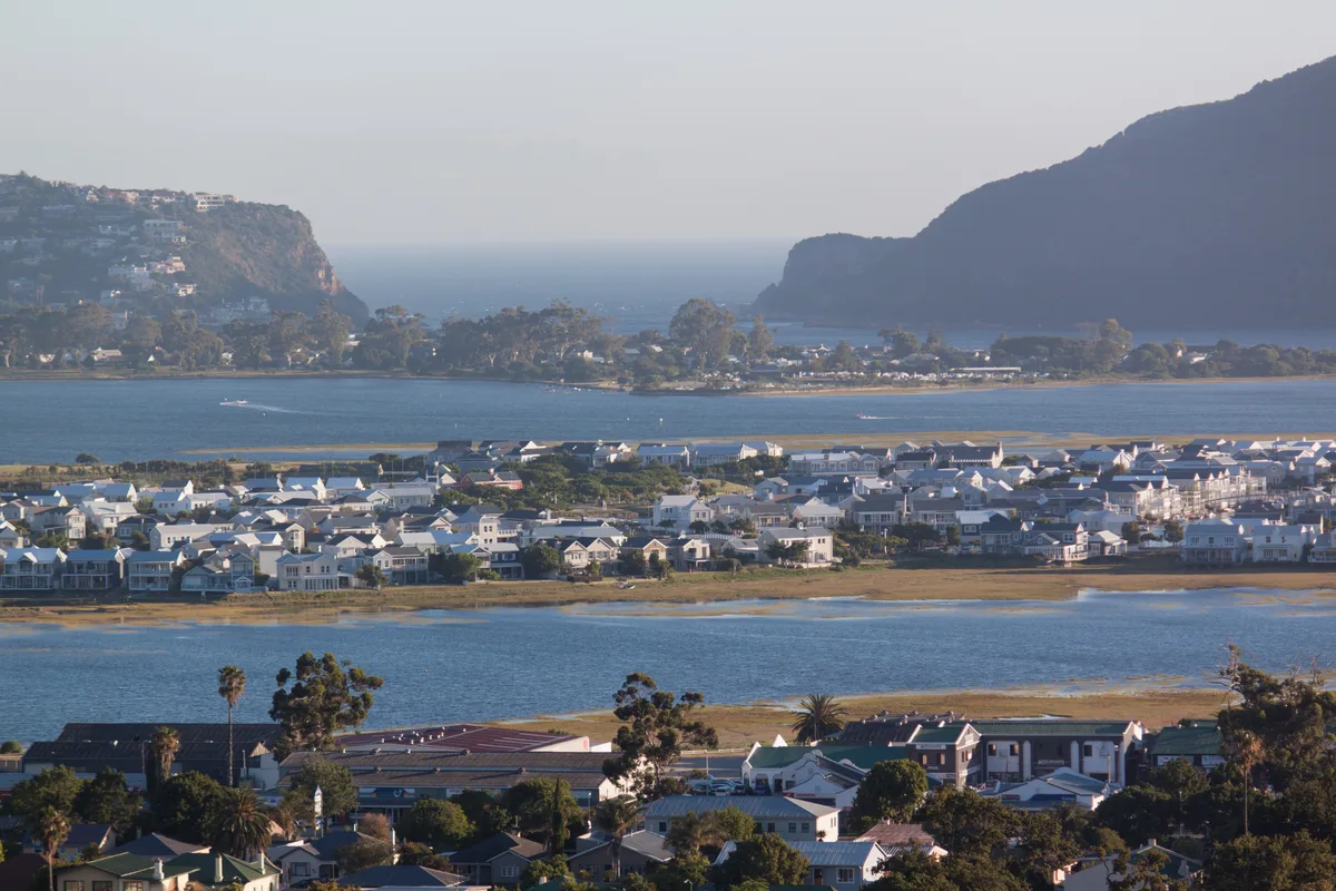 Aerial view across the Knysna Lagoon with Thesen Islands' white canal-estate homes mid-frame and the Heads beyond