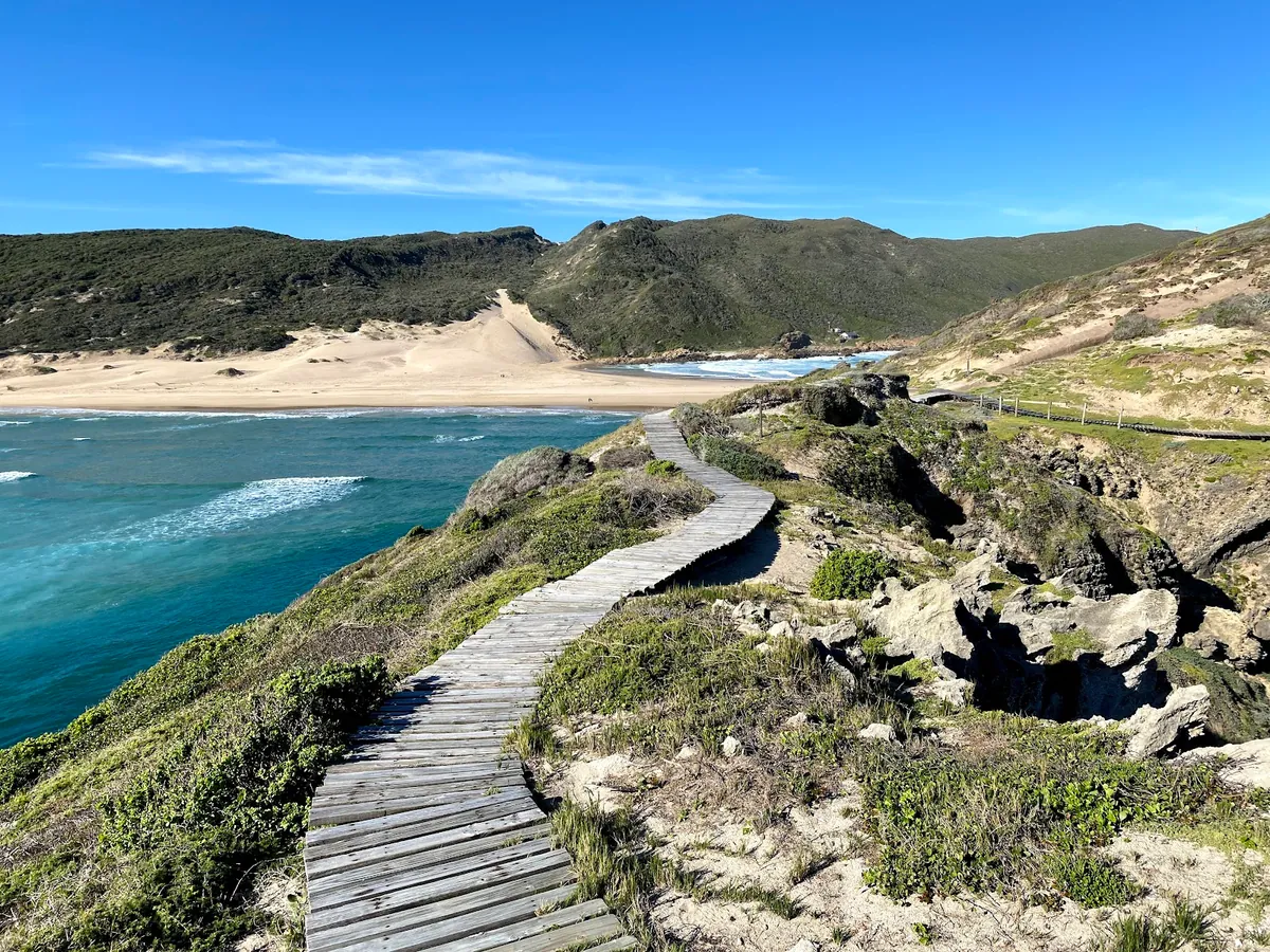 Robberg Nature Reserve boardwalk above the turquoise bay at Plettenberg Bay — timber walkway curving along the cliff, the peninsula and sandy isthmus in the distance
