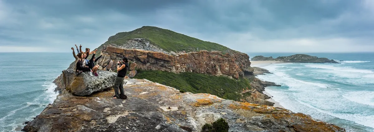 Hikers at a Robberg Nature Reserve viewpoint above the peninsula, with the rocky headland and The Island visible beyond the cliff edge, Plettenberg Bay