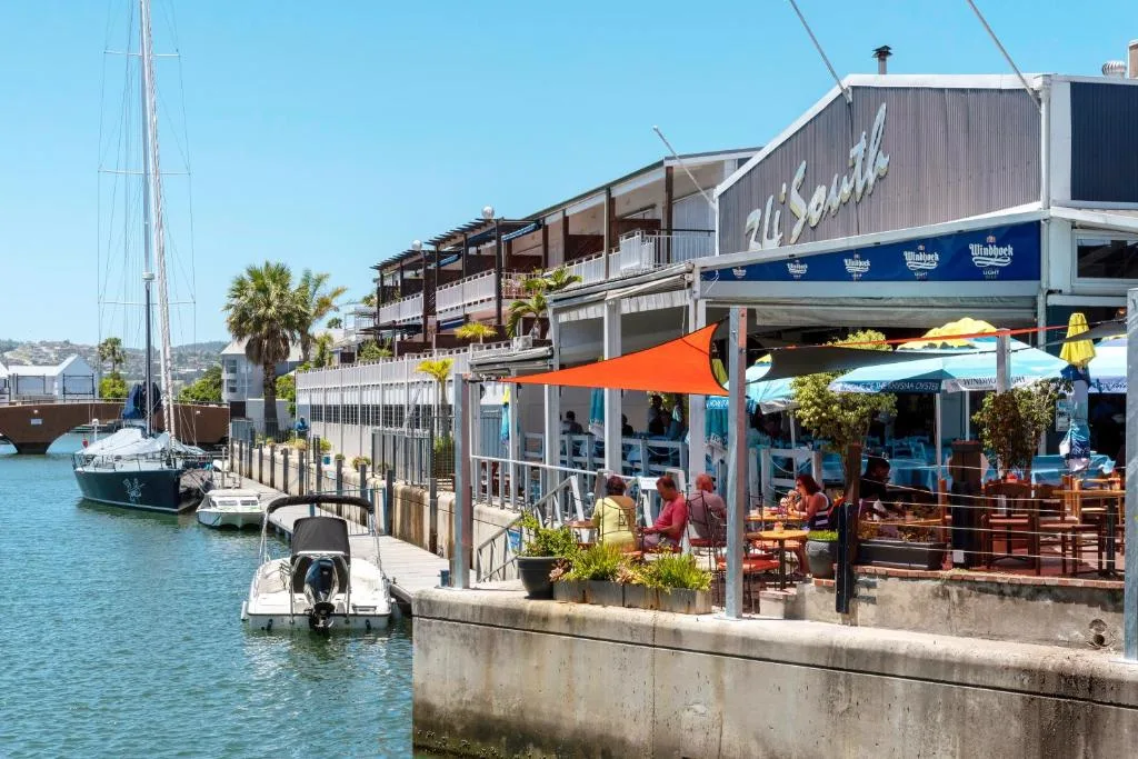 Waterfront harbor with sailboats, restaurants, and coastal buildings under blue sky