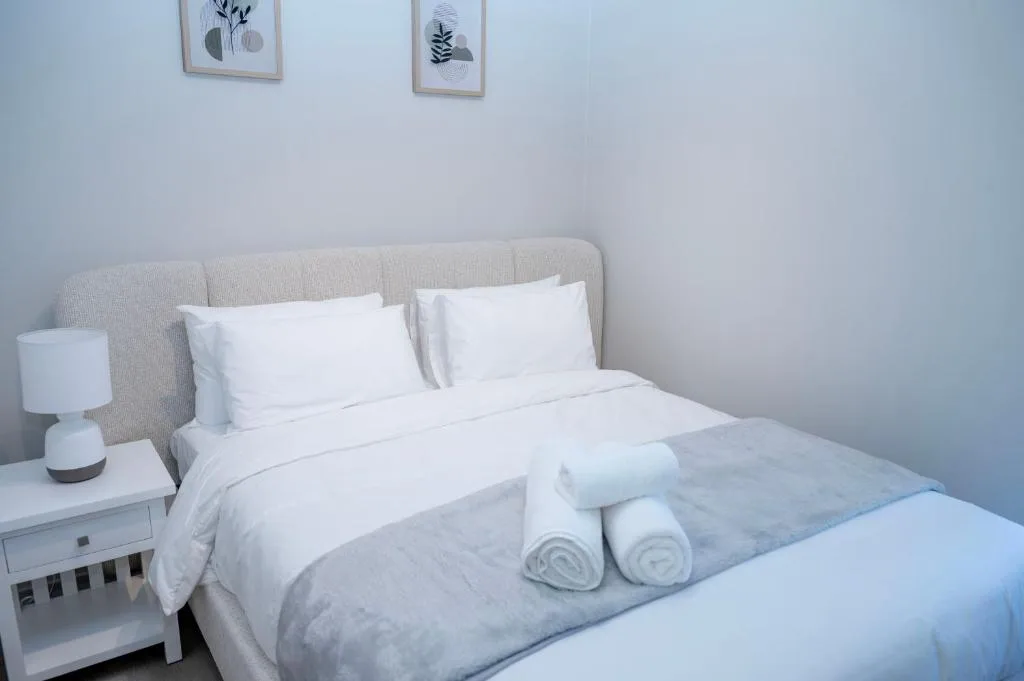 Bright bedroom with queen bed, white pillows, rolled towels, and bedside lamp
