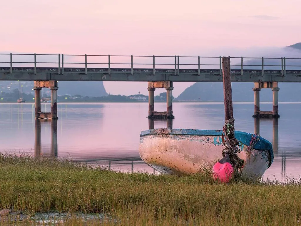 Serene lagoon vista with weathered boat, bridge, and misty mountains at dawn