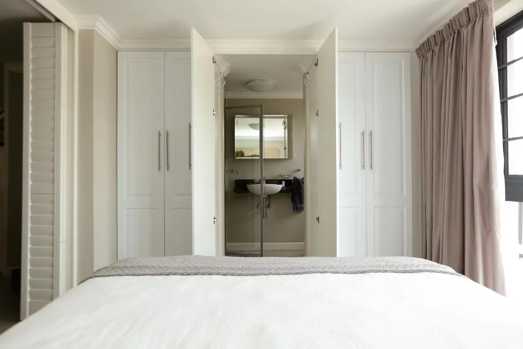 Modern bedroom with white bed and ensuite bathroom visible through glass doors