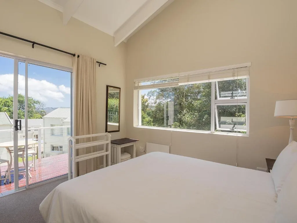 Bright bedroom with white bed, large windows overlooking garden and deck area