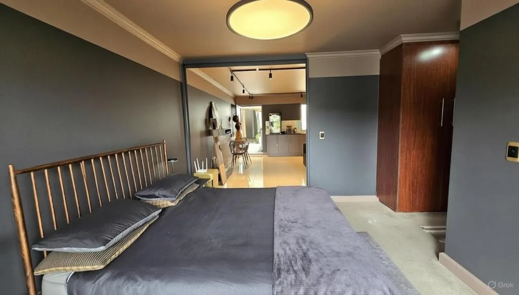 Modern bedroom with dark bed, wooden headboard, open-plan kitchen view beyond