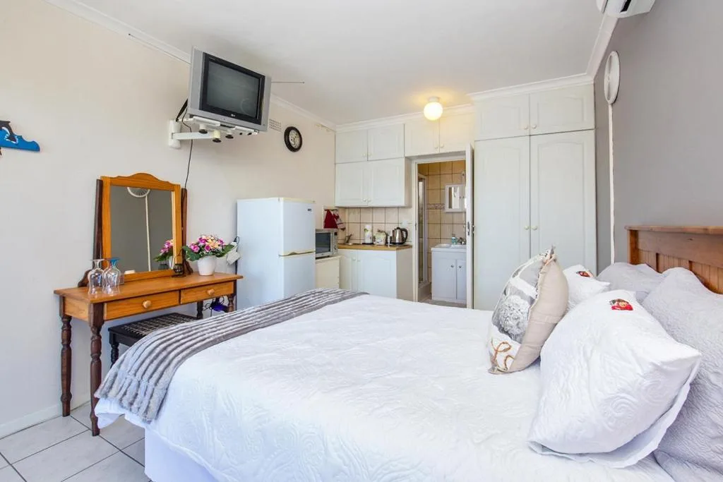 Bright studio bedroom with white bed, wooden dresser, kitchenette, and TV