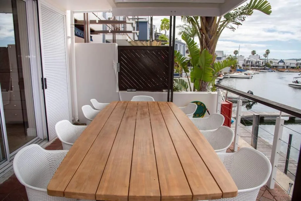 Wooden dining table with white chairs on waterfront deck overlooking lagoon