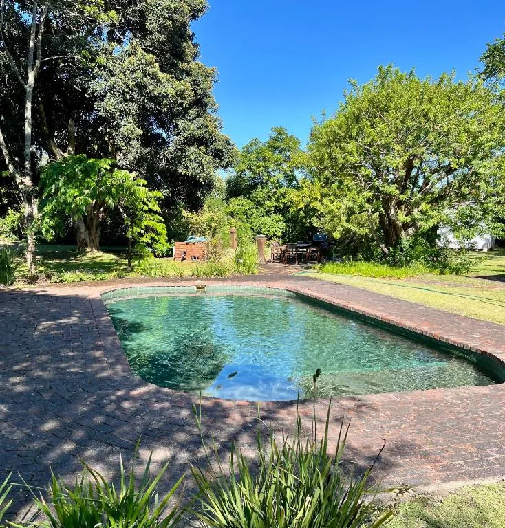 Clear turquoise swimming pool surrounded by lush green trees and brick patio