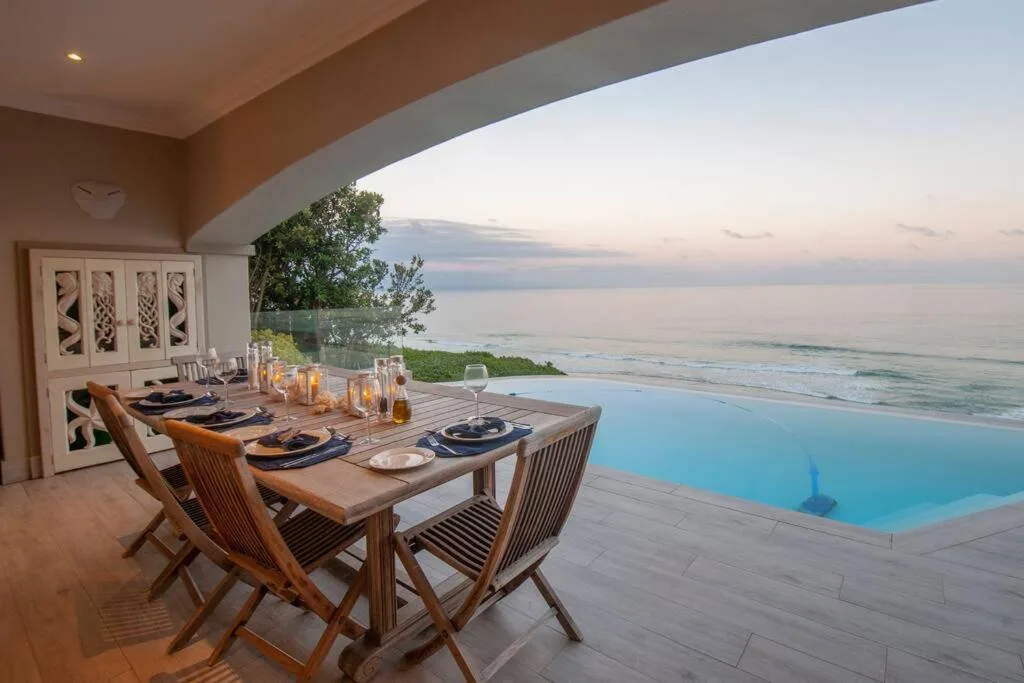 Coastal dining area with ocean view, infinity pool, and set table at sunset