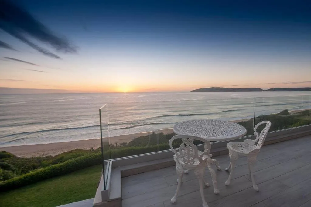 Oceanfront deck with white furniture overlooking beach at sunset