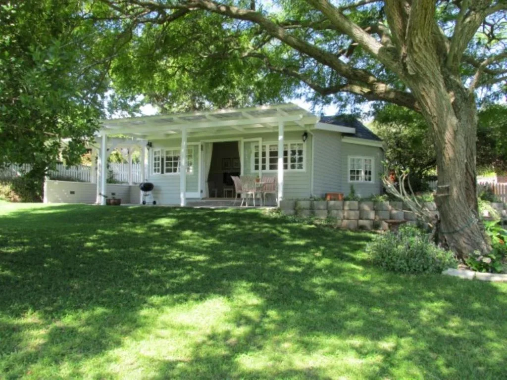 Light green cottage with white pergola and lush green lawn under large tree