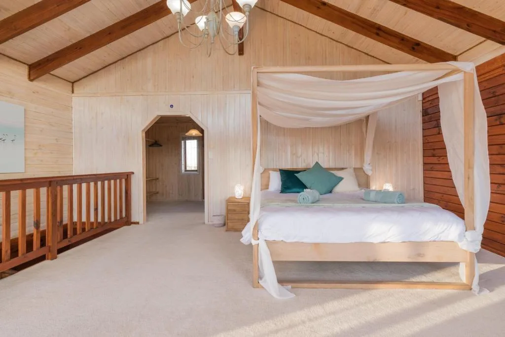 Spacious bedroom with four-poster bed, white drapes, wooden beams, and natural light
