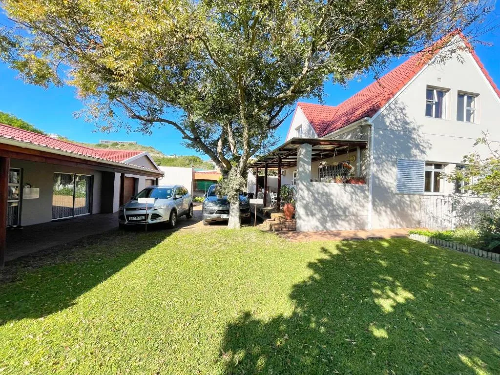 White cottage with red roof, lush green lawn, mature tree, parked cars