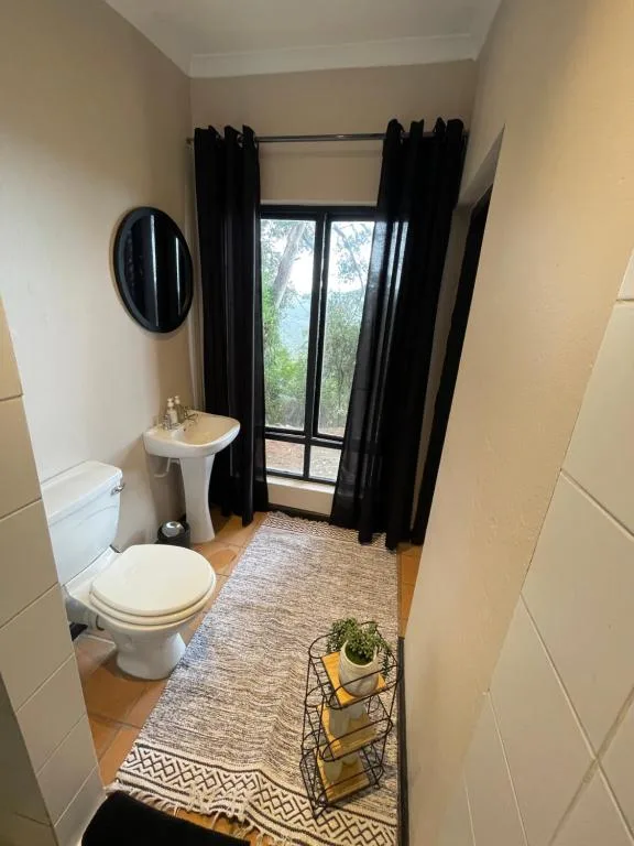 Compact bathroom with toilet, pedestal sink, round mirror, and garden view through window