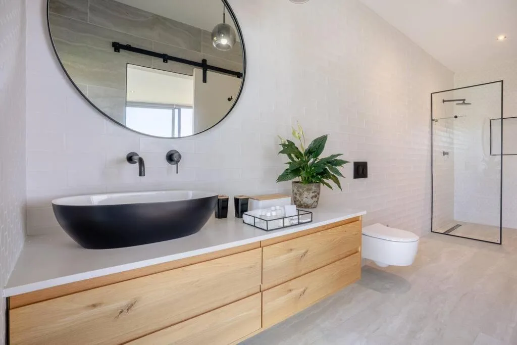 Modern bathroom with black vessel sink, round mirror, wooden vanity, and shower area