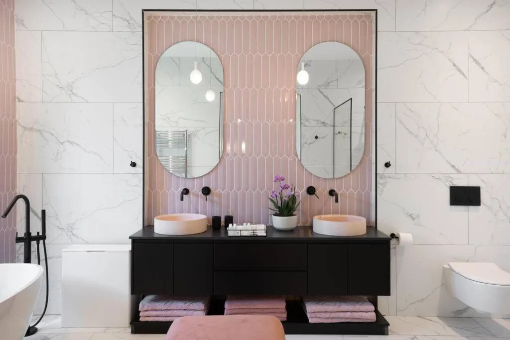Modern bathroom with dual vanities, oval mirrors, marble tiles, and pink accents