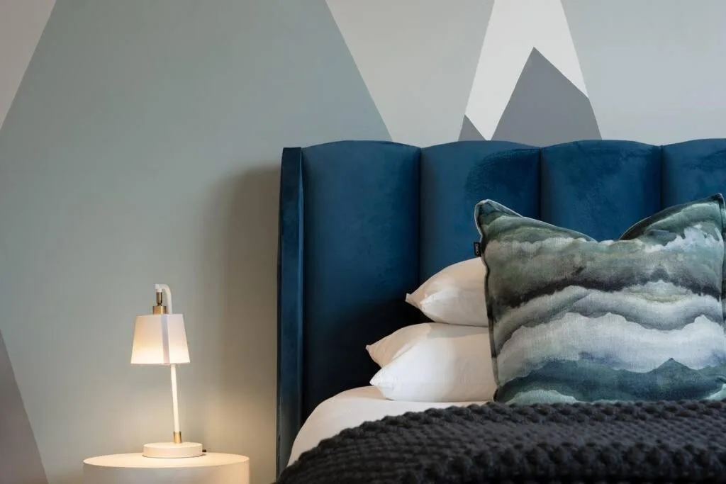 Modern bedroom with blue velvet headboard, layered pillows, and bedside lamp