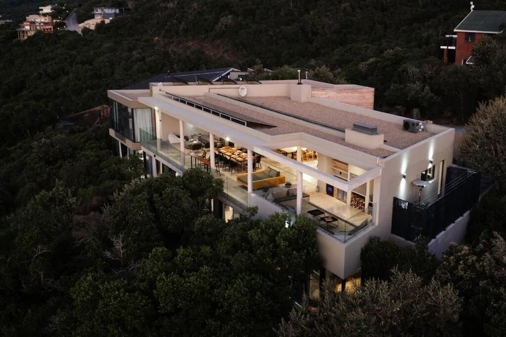 Modern white house nestled in forested landscape at dusk with warm interior lighting