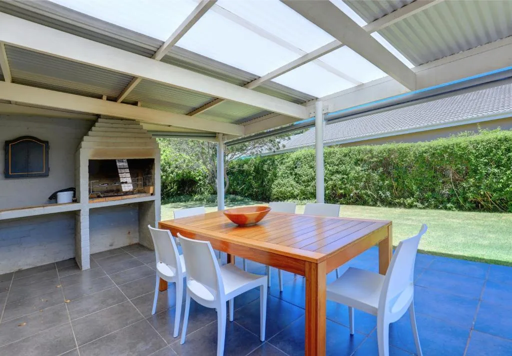 Covered patio with wooden dining table, white chairs, and built-in braai area