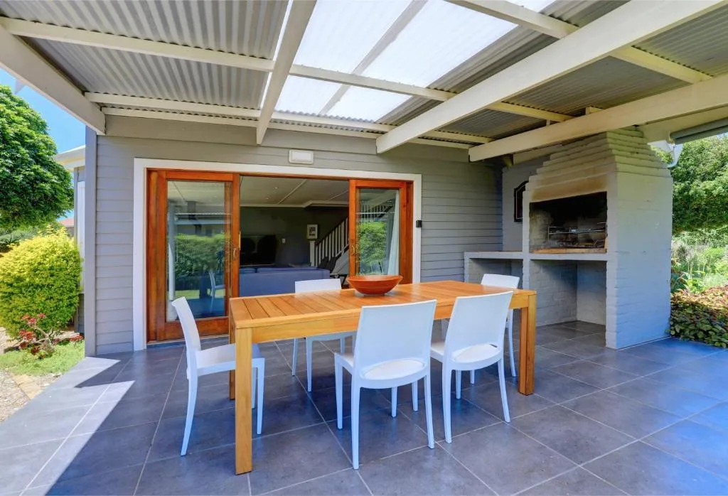 Covered patio with wooden dining table, white chairs, and braai area