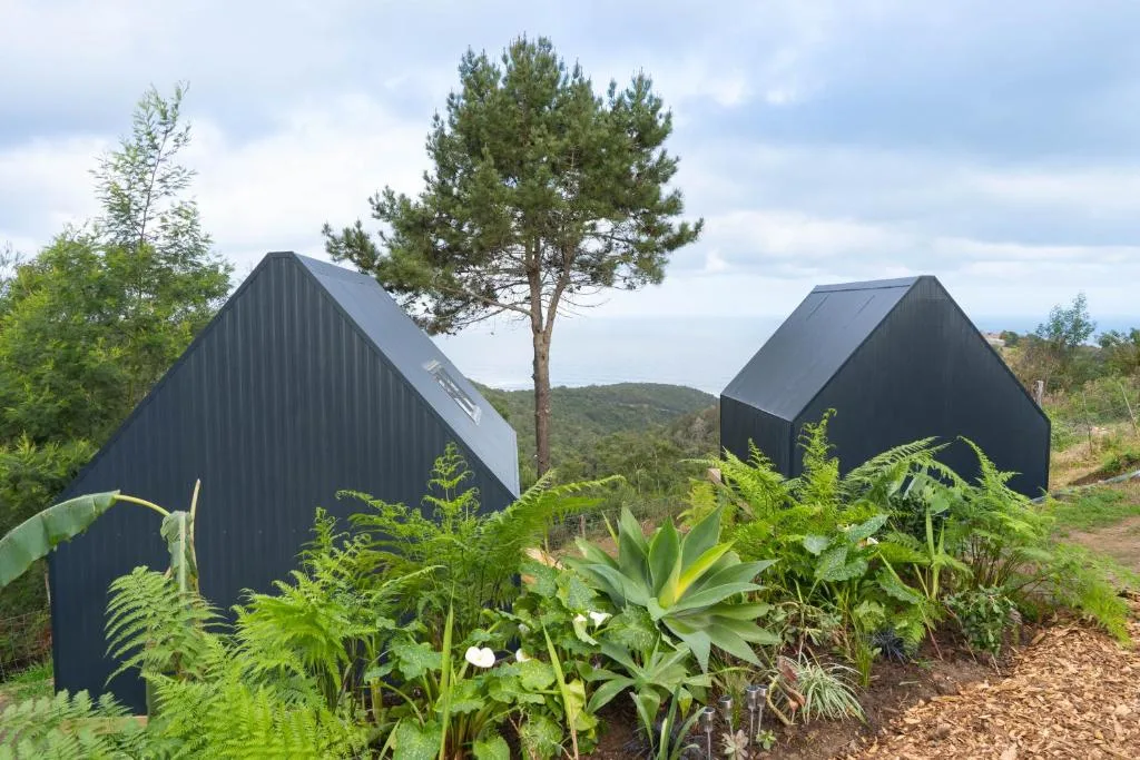 Two black A-frame glamping structures nestled in lush garden landscape with mountains