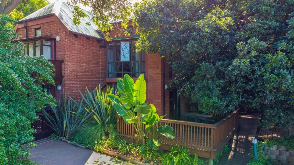 Red brick house with wooden deck surrounded by lush green vegetation and tropical plants