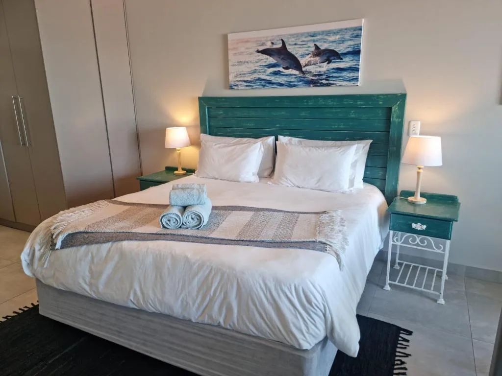 Master bedroom with teal headboard, white bedding, rolled towels, and ocean artwork
